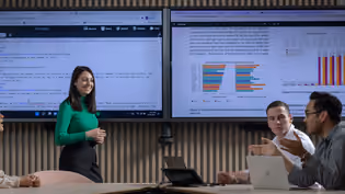Four Business IT students collaborate in a meeting room with two large screens showing code and data visualisations.