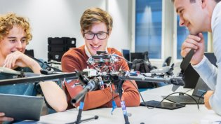Three students collaborating on a robotics project, examining a large drone with exposed wiring and components in a modern laboratory setting. One holds a tablet, while the others observe and discuss the design with interest.