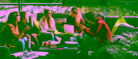 Five students sit on a bench with laptops, notebooks and coffee, having a relaxed group discussion in front of a large window overlooking greenery