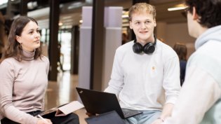 Three students are sitting in an open study space having a conversation. One student has a laptop on their lap and headphones around their neck. Another student is holding a notebook and a pen.