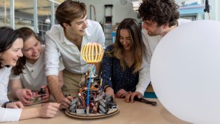 Five students work together on an innovative technical project in a makerspace. They smile and look enthusiastically at a home-made robot with an illuminated, spherical structure and complex wiring. On the table are tools and electronic components. To the right of the robot is a large white balloon. The environment has a creative and technical feel with workbenches and equipment in the background.